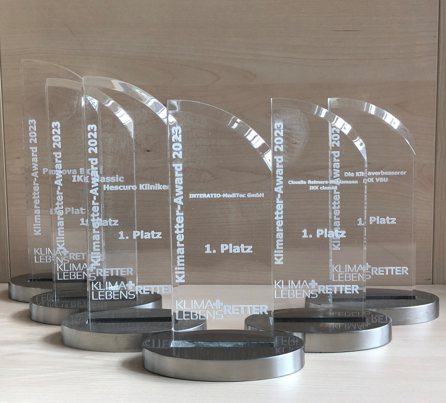 Sechs Trophäen aus Glas stehen auf einem Holztisch, darauf stehen die Namen der Gewinnenden, sowie das Logo des Projekts „Klimaretter-Lebensretter“ und der Text „Klimaretter-Award“.