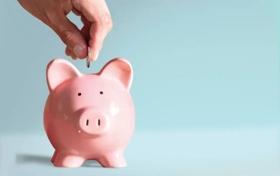 Ein Sparschwein vor blauem Hintergrund. Eine Hand steckt eine Münze in das Sparschwein.