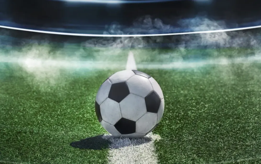 Ein Fußball liegt auf dem Anstoßpunkt eines Spielfelds in einem beleuchteten Stadion unter Flutlicht, im Hintergrund Nebelschwaden. 