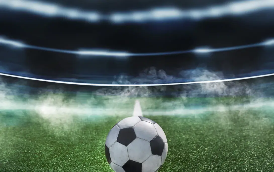 Ein Fußball liegt auf dem Anstoßpunkt eines Spielfelds in einem beleuchteten Stadion unter Flutlicht, im Hintergrund Nebelschwaden. 