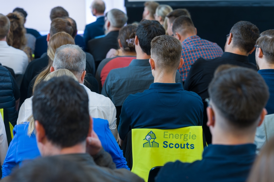 Menschen von hinten, sitzen dicht nebeneinander in einem Seminarraum. Eine Person in der Mitte trägt eine Weste mit der Aufschrift "Energie Scouts".