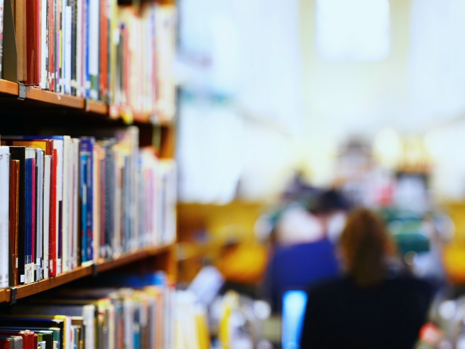 Eine Reihe Bücher in einer Bibliothek, im Hintergrund sind Lesende an Tischen zu sehen.