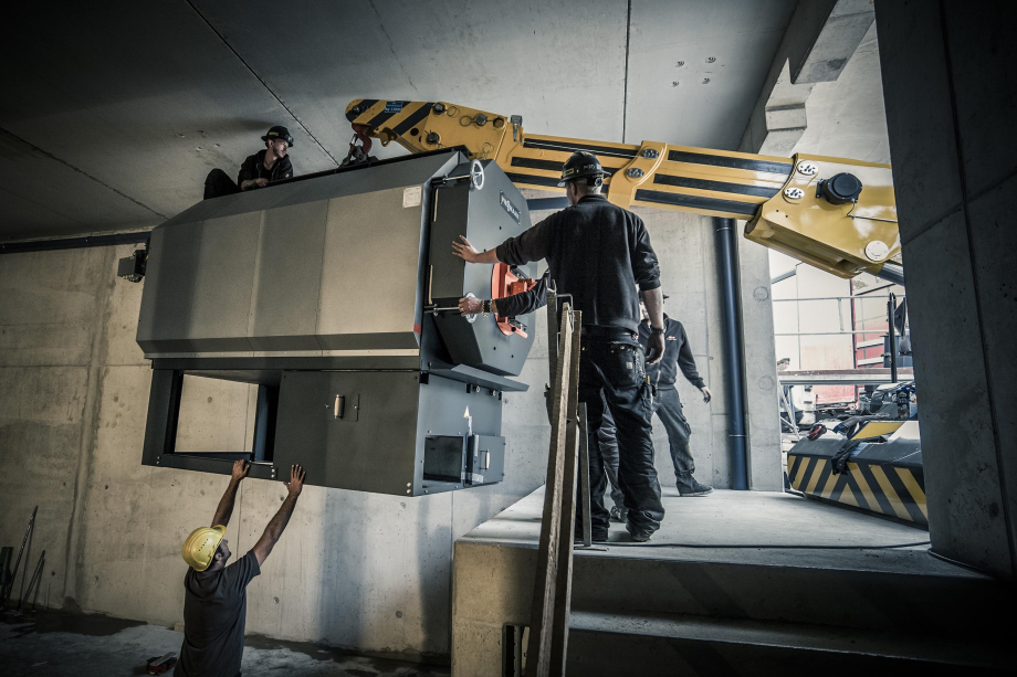 Ein großes Kesselgerät wird mit einem Kran in eine Halle transportiert, Menschen mit Helm dabei