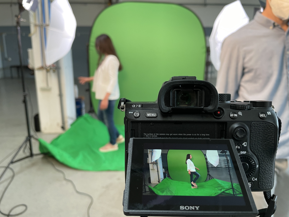 Auf einem Kameradisplay sieht man eine Frau vor einem Greenscreen. Im Hintergrund steht diese Frau vor dem Greenscreen.