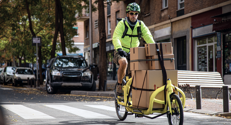 Fahrer mit Warnjacke transportiert Pakete auf einem Lastenrad durch eine Stadtstraße.