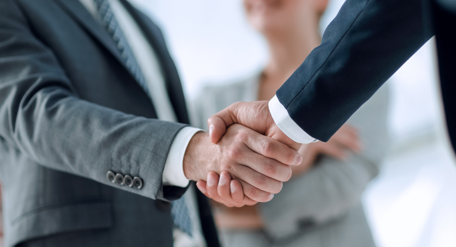 Zwei Personen in Businesskleidung geben sich zur Begrüßung die Hand, eine weitere Person steht im Hintergrund.