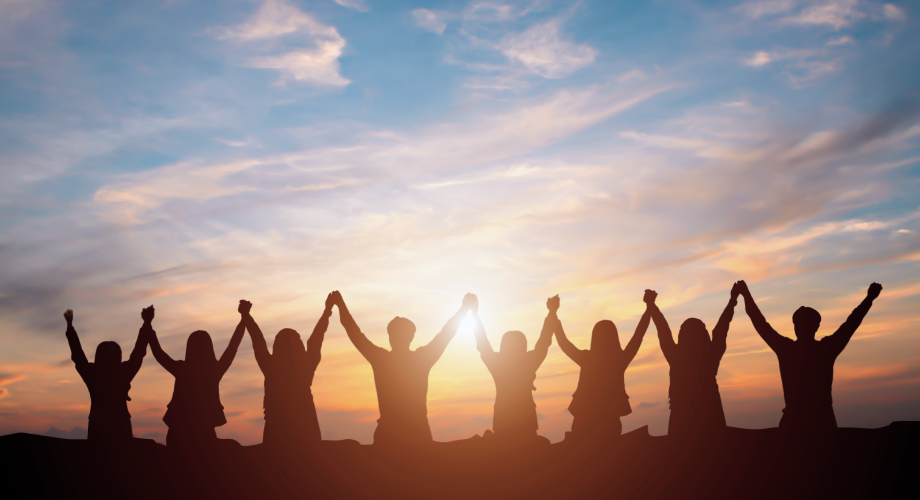 Menschen stehen im Sonnenuntergang nebeneinander und halten einander die erhobenen Hände.