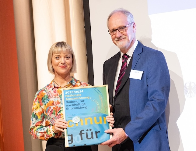 Nadja Kulikowa (links, WWF) und Dr. Klaus Grosfeld (rechts, REKLIM) stehen auf der Bühne und halten die Urkunde für die „Nationale Auszeichnung – Bildung für nachhaltige Entwicklung“ in den Händen. Beide blicken freundlich in die Kamera.