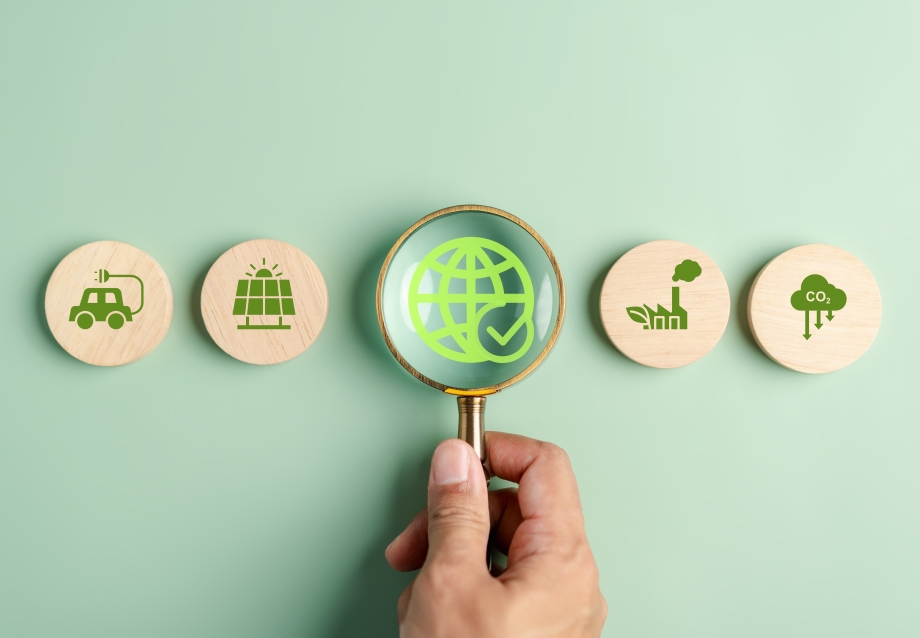 Eine Lupe ist auf runde Holzbausteine gerichtet. Auf ihnen sind in grün Symbole zu sehen: Ein Elektroauto, eine Photovoltaikanlage, eine Weltkugel, eine Fabrik, und eine Wolke, auf der CO2 steht.