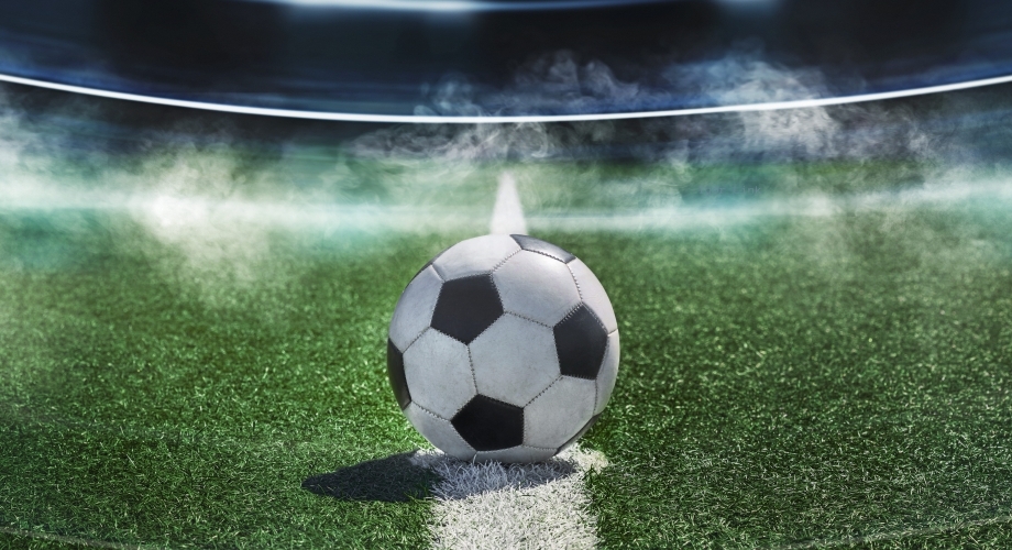Ein Fußball liegt auf dem Anstoßpunkt eines Spielfelds in einem beleuchteten Stadion unter Flutlicht, im Hintergrund Nebelschwaden. 