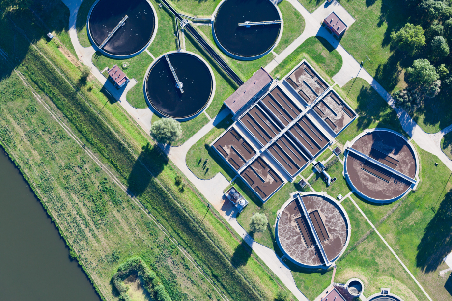 Luftaufnahme einer Kläranlage mit runden und eckigen Becken, umgeben von Rasenflächen. In der linkenEcke durchschneidet ein Wasserlauf das Bild. Rechts oben sind einige Bäume zu sehen. 