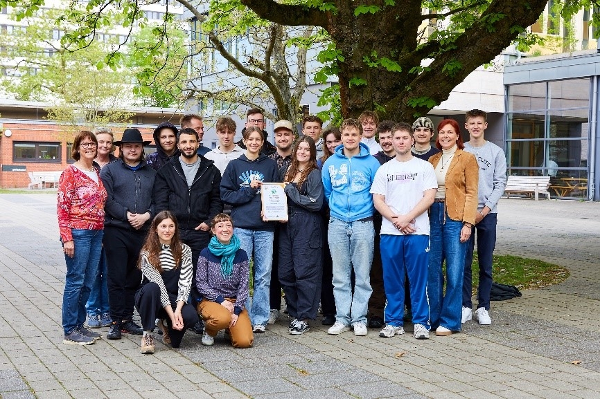 Gruppe Jugendlicher und Erwachsener steht unter einem Baum im Schulhof, eine Person hält eine Urkunde.