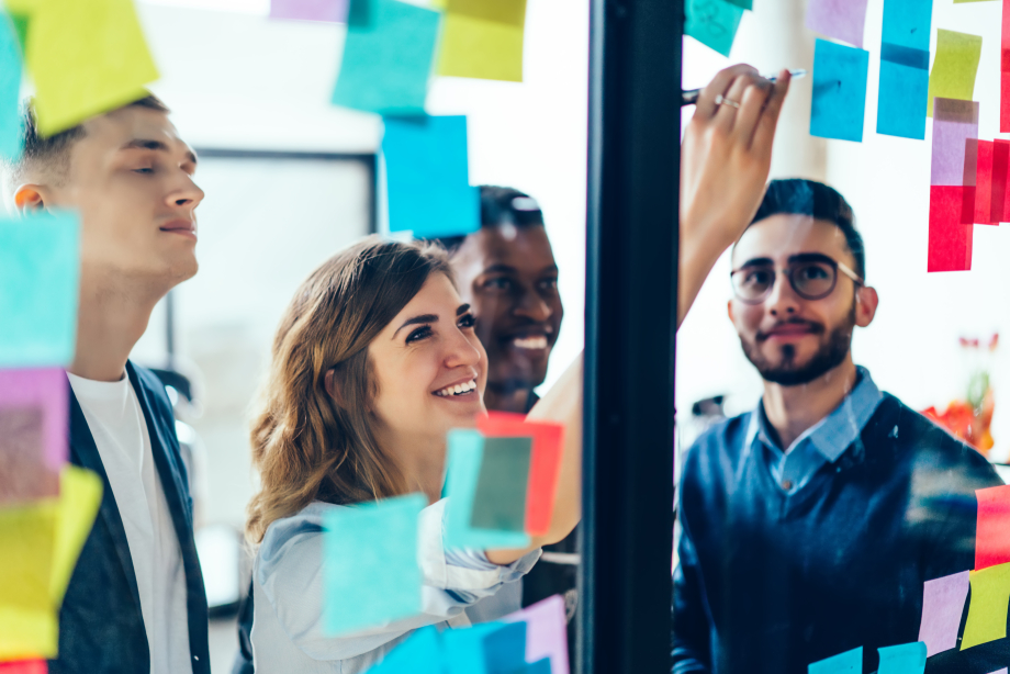 Eine Frau klebt ein beschriebenes Post-it an eine Wand. Sie ist umringt von anderen Workshop-Teilnehmenden.