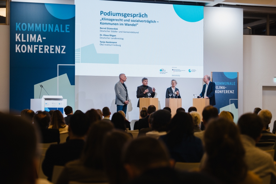 Drei Männer und eine Frau stehen auf einer Bühne an Stehlen und diskutieren miteinander.