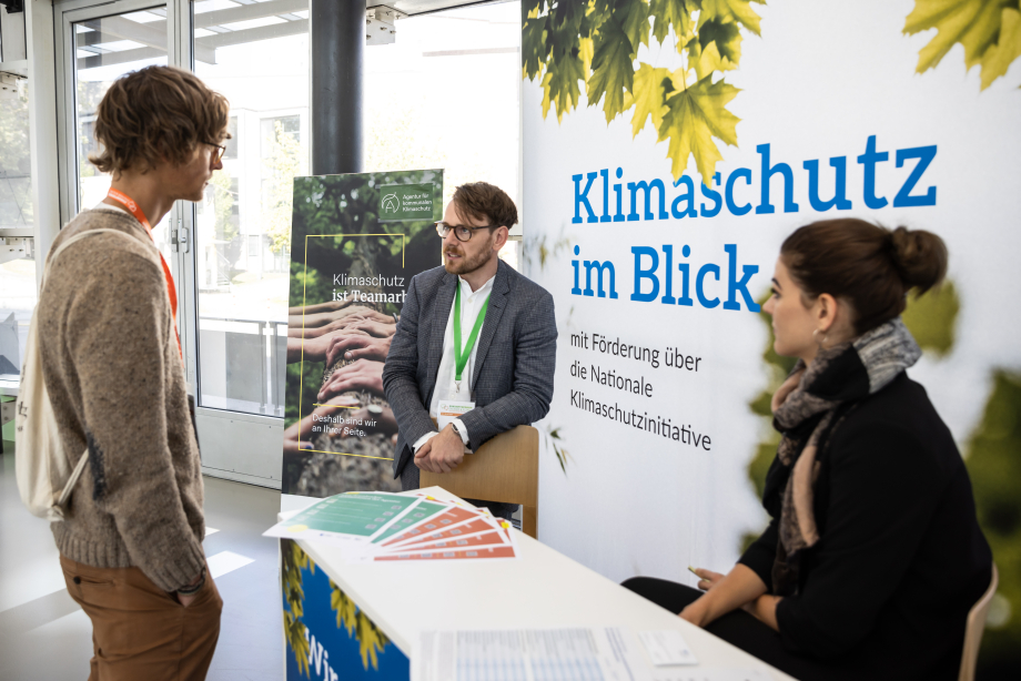 Zwei Mitarbeitende der Agentur für kommunalen Klimaschutz unterhalten sich an ihrem Informationsstand mit einer interessierten Person.