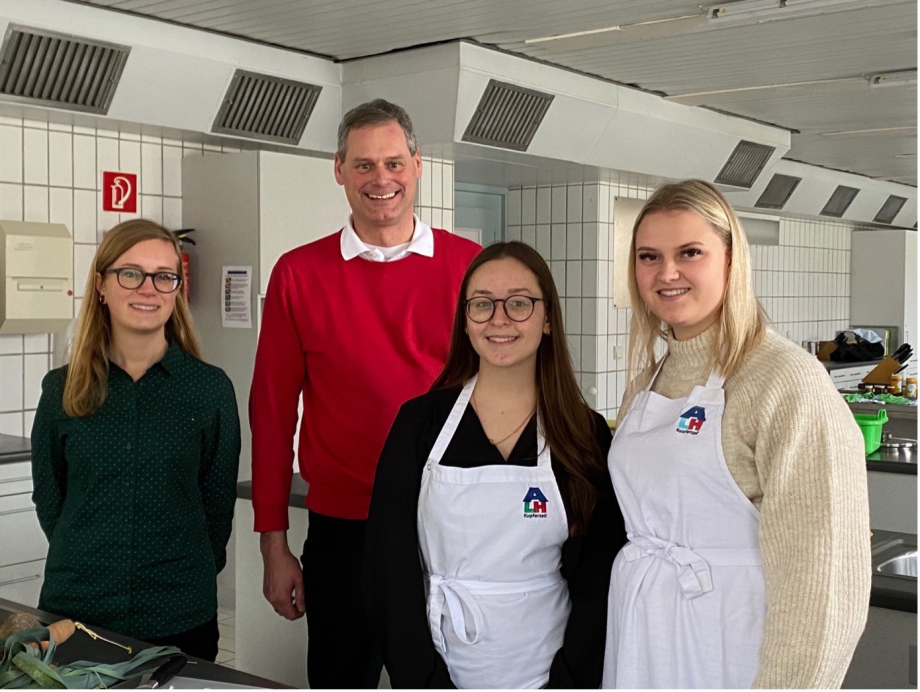 Vier Personen stehen in einer Großküche, zwei von ihnen haben weiße Schürzen an.