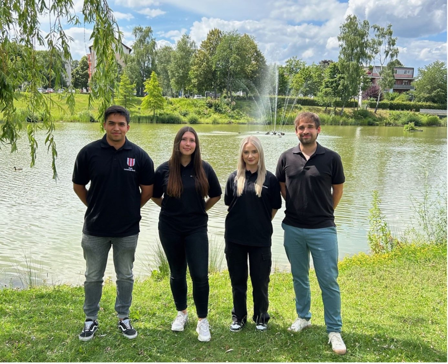 Vier Personen stehen vor einem Teich mit Springbrunnen im Grünen.