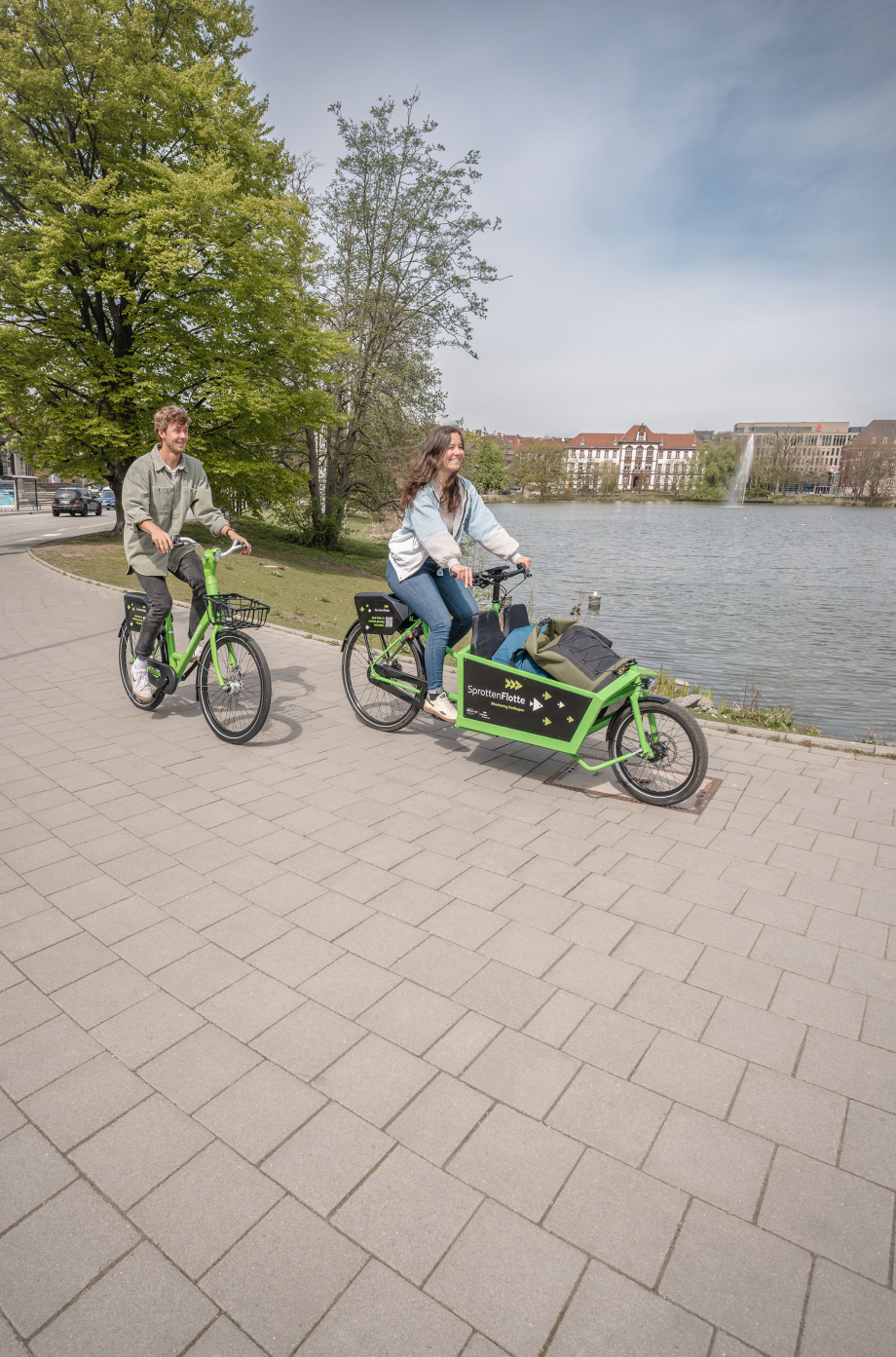 Zwei Personen fahren auf Fahrädern (einem Lastenrad und einem normales Rad) an einem Flussufer entlang.