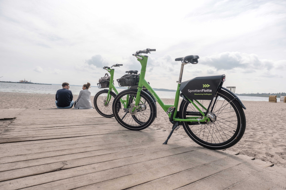 Zwei grüne Bike-Sharing-Fahrräder der SprottenFlotte stehen an einem Sandstrand.
