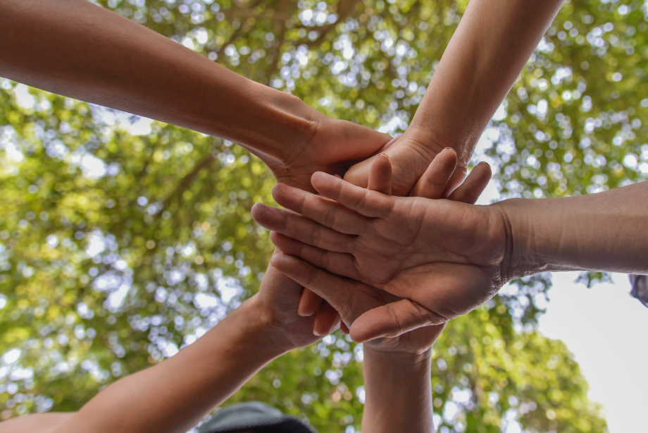 Mehrere Menschen halten ihre Hände in einem Kreis übereinander als Symbol des Zusammenhalts. Im Hintergrund sind Baumkronen zu erkennen. 