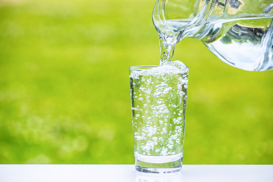 In ein Glas wird Wasser aus einem Krug eingegossen. Im Hintergrund ist eine Wiese zu sehen. 