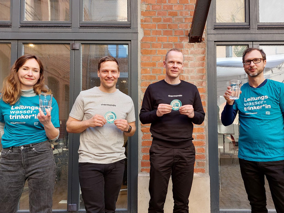 Das Foto zeigt eine Frau und drei Männer vor einer Hauswand, die den Aufkleber „Leitungswasserfreundliches Unternehmen“ des Vereins a tip: tap in den Händen halten.