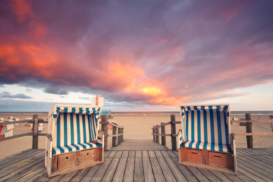 Das Foto zeigt zwei Strandkörbe. Im Hintergrund sieht man den Strand und einen Sonnenuntergang.