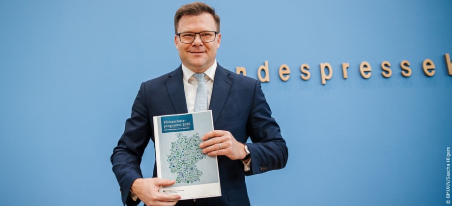Bundesumweltminister Carsten Schneider steht vor blauem Hintergrund und hält das Klimaschutzprogramm 2026 in den Händen.