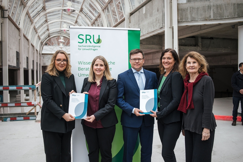 Fünf Personen stehen vor einem SRU-Banner in einem Rohbau und halten Broschüren in den Händen.