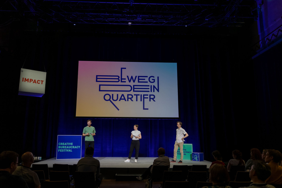 Drei Personen stehen auf einer Bühne. Im Hintergrund ein Bildschirm auf dem „Beweg dein Quartier“ steht.