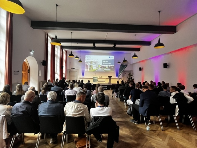 In einem Saal sitzen zahlreiche Menschen vor einer Leinwand mit einer Präsentation.