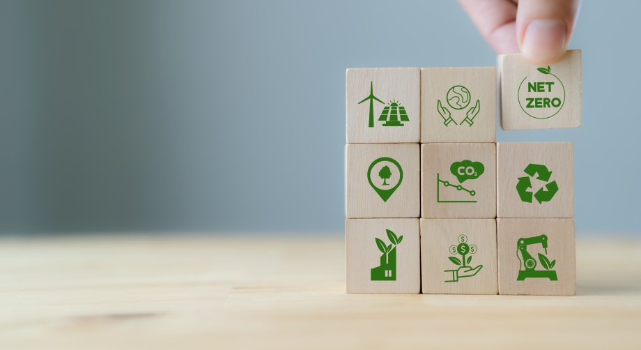 Holzbausteine mit grünen Klimaschutzsymbolen, wie eine Pyramide angeordnet, der oberste Baustein mit der Aufschrift 'net Zero' wird von einer Hand auf die Spitze gesetzt. 