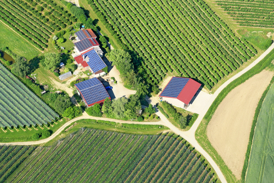 Von oben: landwirtschaftliche Felder mit drei Gebäuden in der Mitte auf denen Photovoltaikanlagen installiert sind