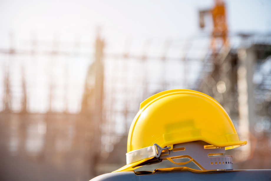 Auf dem Foto zu sehen ist die Seitenansicht eines gelben Baustellenhelms. Im Hintergrund verschwommen zu erkennen ist ein Metallgerüst für das Gießen von Beton.