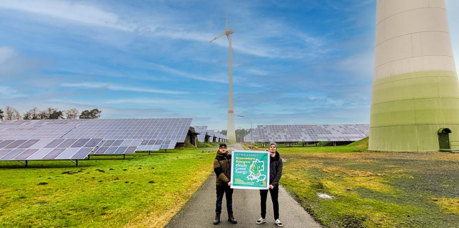 Zwei Personen stehen vor Windrädern und Photovoltaikanlagen und halten ein Plakat mit dem Keyvisual von #StudyGreenEnergy