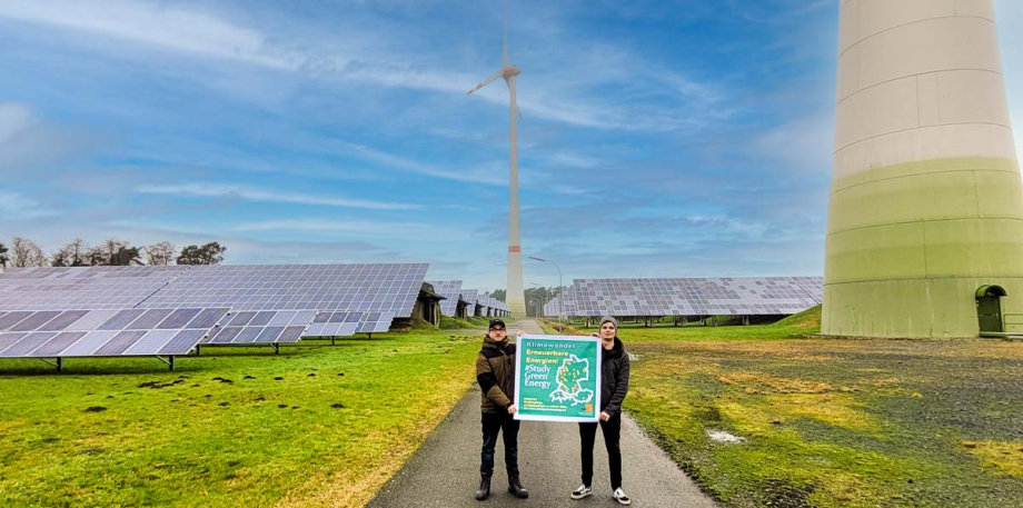 Zwei Personen stehen vor Windrädern und Photovoltaikanlagen und halten ein Plakat mit dem Keyvisual von #StudyGreenEnergy