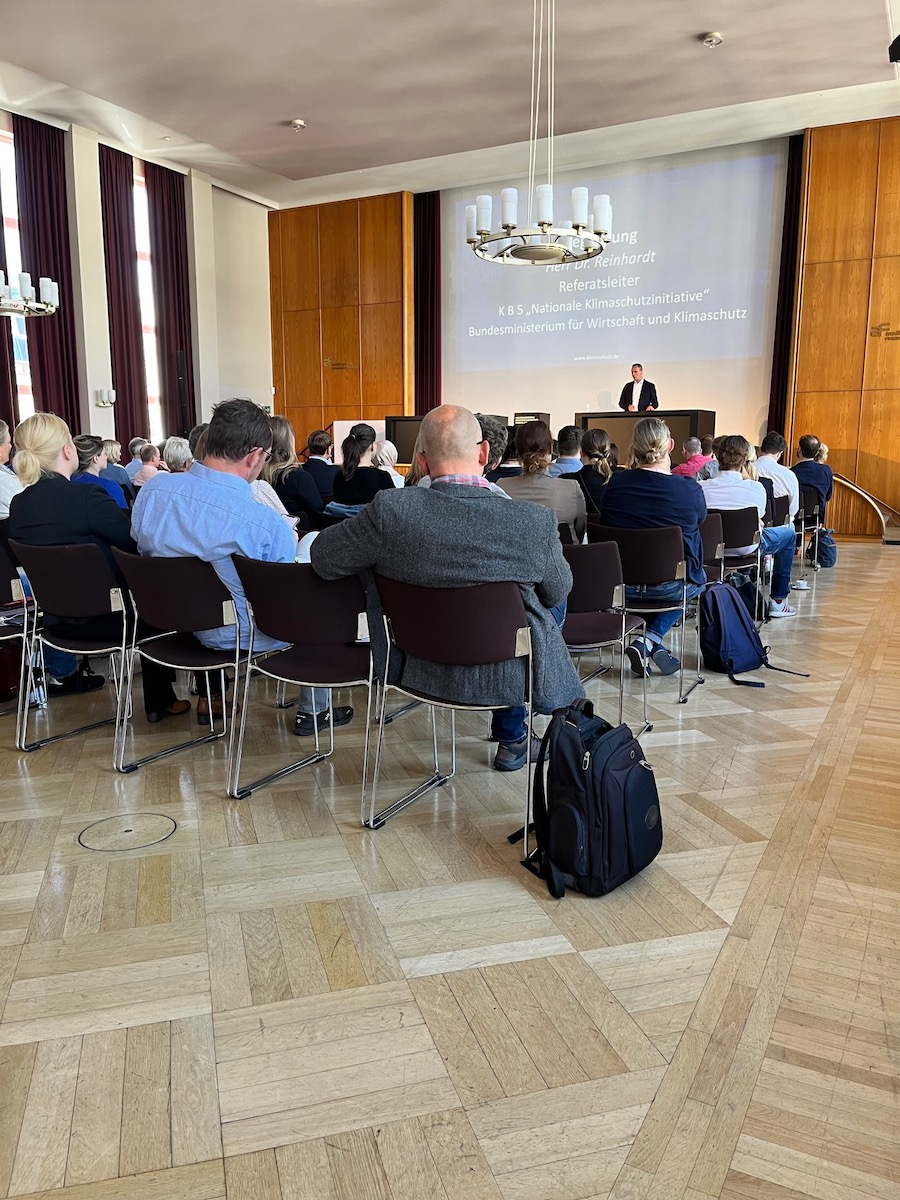 Im Auditorium Friedrichstraße hält Herr Dr. Reinhardt, Referatsleiter KB5 „Nationale Klimaschutzinitiative“ im Bundesministerium für Wirtschaft und Klimaschutz, eine Grußrede vor Publikum. Herr Reinhardt steht auf der Bühne am Tisch und hat die Hände zu einer einladenden Geste geöffnet.