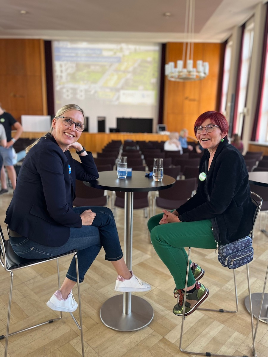 Zwei Teilnehmerinnen des Vernetzungstreffens für innovative Klimaschutzprojekte sitzen gemeinsam am Tisch und lächeln uns an. Im Hintergrund sieht man den holzgetäfelten Saal des Auditoriums Friedrichstraße.
