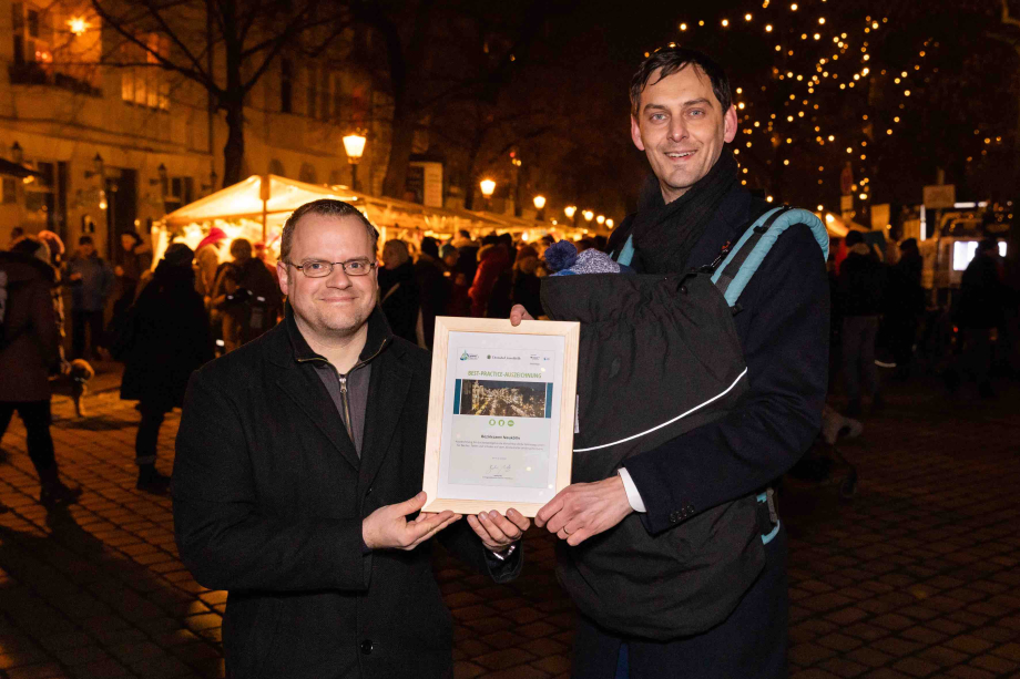 Thomas Fischer, Leiter Kreislaufwirtschaft Deutsche Umwelthilfe und Martin Hikel, Bezirksbürgermeister von Berlin-Neukölln, stehen nebeneinander und halten eine gerahmte Urkunde in der Hand. Martin Hikel hat ein Baby in einem Tragerucksack vor der Brust. Im Hintergrund ist der Alt-Rixdorfer Weihnachtsmarkt zu sehen. 