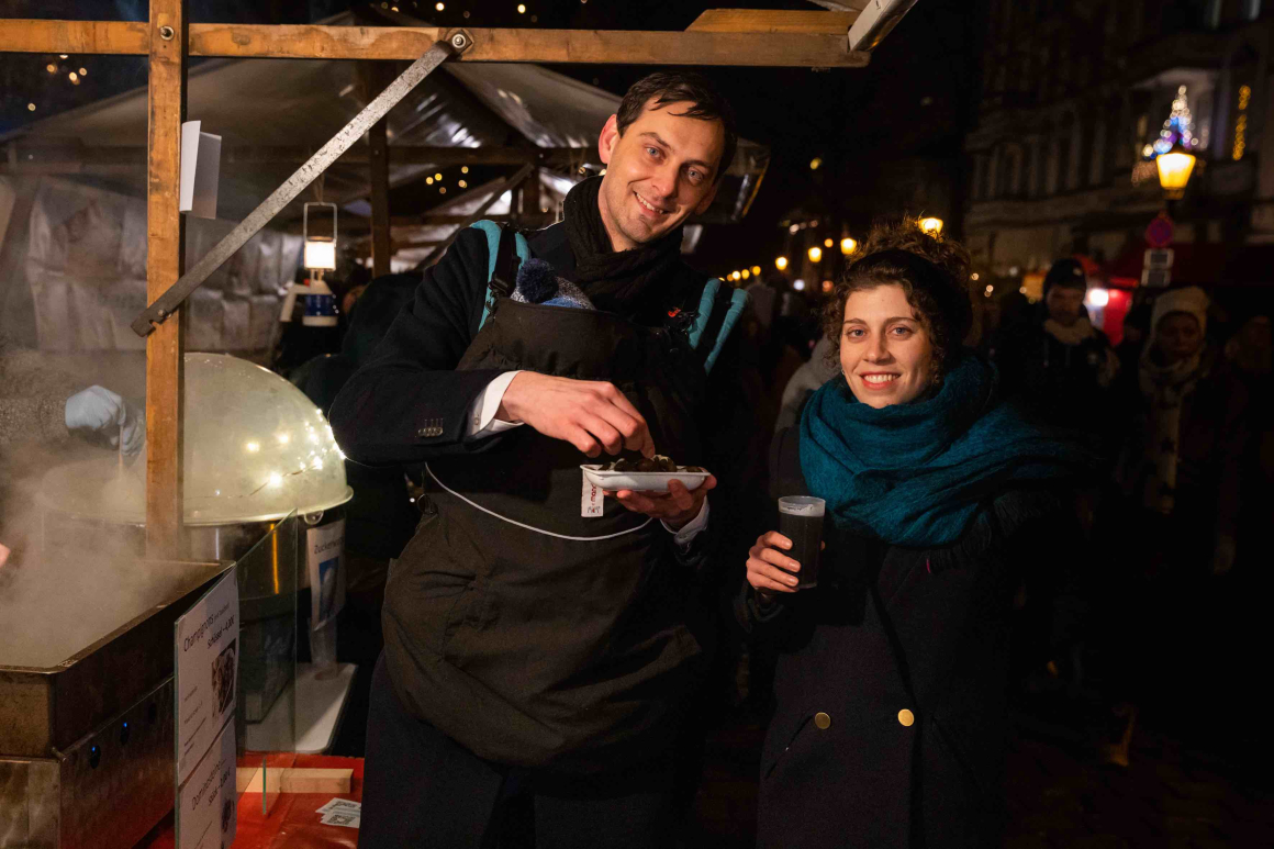 Der Bezirksbürgermeister von Berlin-Neukölln, Martin Hikel, und Elena Schägg, Senior Expert Kreislaufwirtschaft der Deutschen Umwelthilfe, stehen nebeneinander an ei-nem Stand auf dem Alt-Rixdorfer Weihnachtsmarkt. Martin Hikel hat ein Baby in einem Tragerucksack vor der Brust und hält eine Mehrwegschale mit Essen in der Hand. Elena Schägg hält einen Mehrwegbecher mit Glühwein.