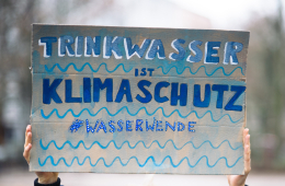 Das Bild zeigt ein gemaltes Plakat mit der Aufschrift „Trinkwasser ist Kilmaschutz. Hashtag Wasserwende.