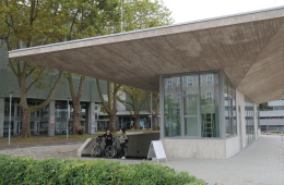 Das Bild zeigt die neue Fahrradstation an der Universität zu Köln und eine Studierende und Mitarbeitende mit ihren Fahrrädern in der Einfahrt des Gebäudes.