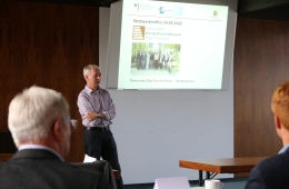 Ein Mann steht vor einer Leinwand, auf der eine Power-Point-Präsentation läuft. Mitglieder des Netzwerks sitzen im Publikum.