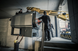 Ein großes Kesselgerät wird mit einem Kran in eine Halle transportiert, Menschen mit Helm dabei