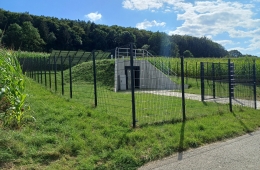 Seitenansicht der Pumpwerks in Niederlistingen. Die Anlage ist umzäunt mit einem schwarzen, hohen Metallzaun. Vor der Anlage eine Rasenfläche. Die Anlage besitzt im Vordergrund Betonwände mit der Eingangstür im Zentrum. Über den Betonwänden metallene Brüstungen. Hinter dem Eingang aus Beton eine hügelähnliche, von Gras bedeckte Erhöhung. Hinter der Anlage befinden sich mehrere Solarpanele, die auf der Wiese aufgestellt sind. Neben der Anlage Maisfelder und im Hintergrund der Beginn eines Waldstücks. 