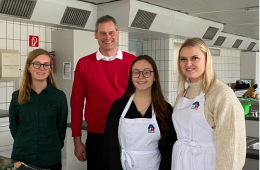 Vier Personen stehen in einer Großküche, zwei von ihnen haben weiße Schürzen an.