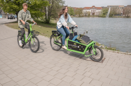 Zwei Personen fahren auf Fahrädern (einem Lastenrad und einem normales Rad) an einem Flussufer entlang.