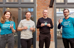 Das Foto zeigt eine Frau und drei Männer vor einer Hauswand, die den Aufkleber „Leitungswasserfreundliches Unternehmen“ des Vereins a tip: tap in den Händen halten.