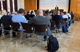 Im Auditorium Friedrichstraße hält Herr Dr. Reinhardt, Referatsleiter KB5 „Nationale Klimaschutzinitiative“ im Bundesministerium für Wirtschaft und Klimaschutz, eine Grußrede vor Publikum. Herr Reinhardt steht auf der Bühne am Tisch und hat die Hände zu einer einladenden Geste geöffnet.