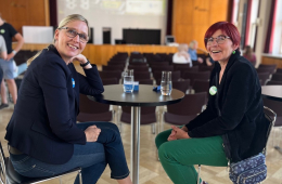 Zwei Teilnehmerinnen des Vernetzungstreffens für innovative Klimaschutzprojekte sitzen gemeinsam am Tisch und lächeln uns an. Im Hintergrund sieht man den holzgetäfelten Saal des Auditoriums Friedrichstraße.